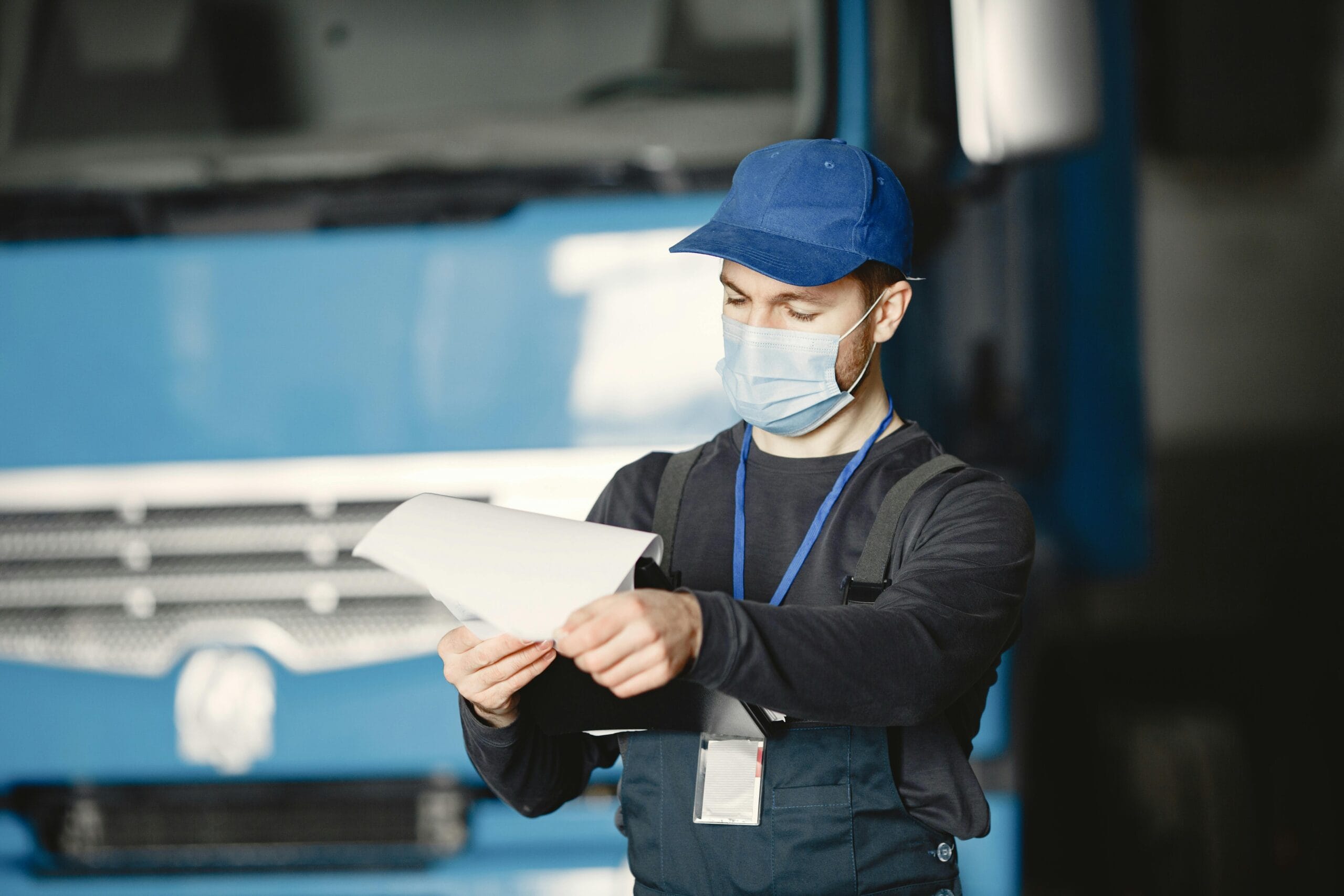 Mechanic wearing blue cap and face mask reviewing documents in auto workshop.