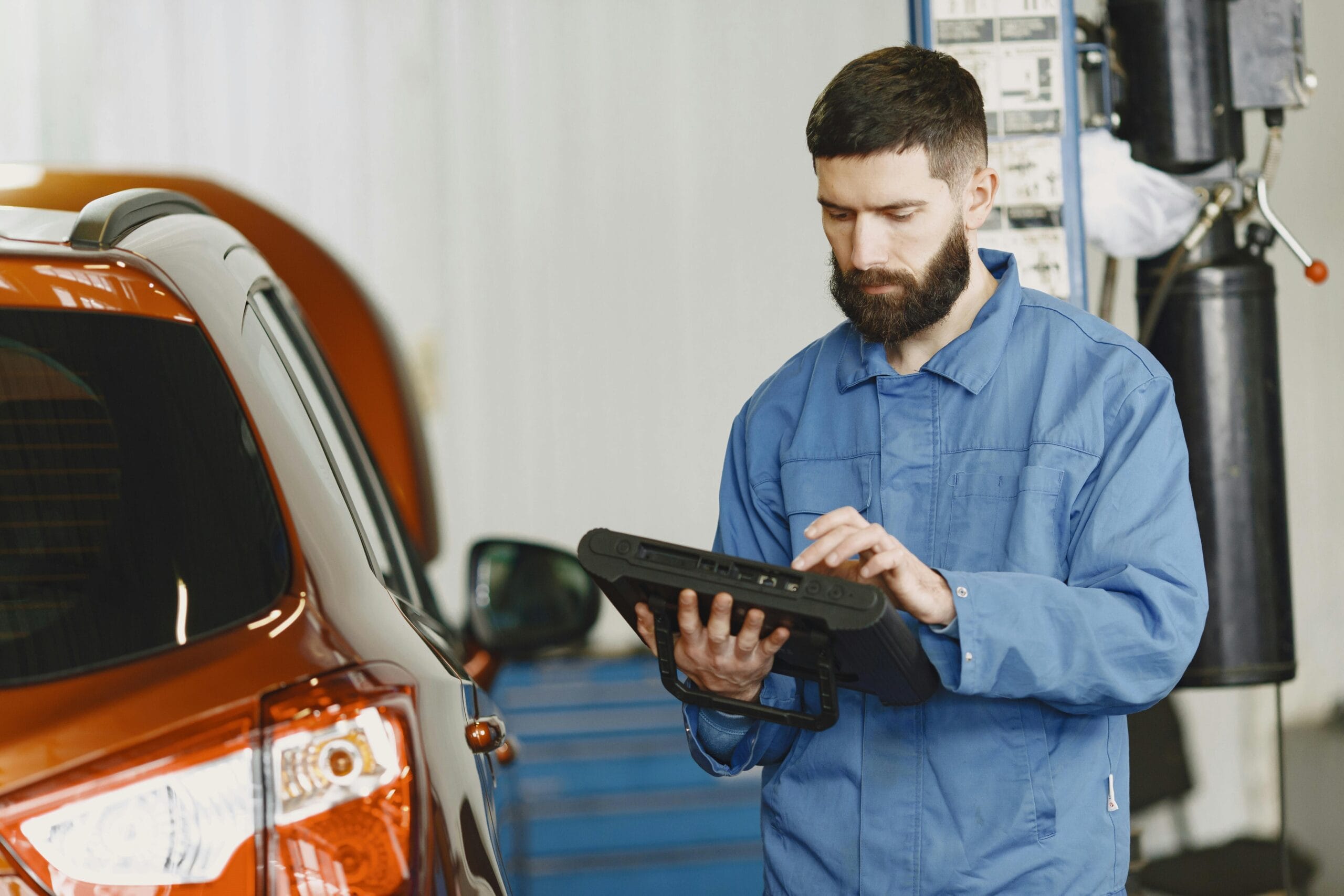 A mechanic in coveralls using a diagnostic tool at an auto repair shop beside a car.